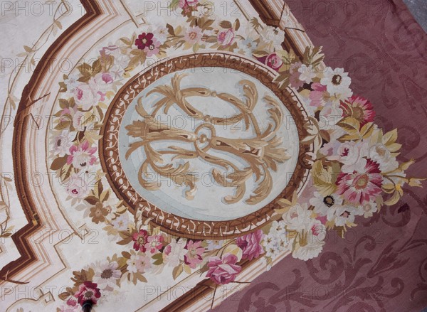 A so-called ceiling tapestry, tapestries, fabrics, tapestries, fabrics, knotted monograms, floral motifs, interior furnishings, weaving. In the center, a rosette surrounded by a floral wreath. Surrounded by rectangular frames with rounded shorter sides and floral garlands. In the corners are medallions with a knotted CKJ monogram. The border varies in color with plant patterns imitating the damask technique. Colors: pink, red, light brown, and olive. Early 19th century, France. Height 681 cm, width 385 cm., A decorative circular medallion with ornate swirling leaf motifs in beige and gold against a pale blue center, surrounded by a brown patterned border and a garland of roses and other flowers in shades of pink, white, cream, and muted yellow with green and brown leaves, set on a background that transitions from a light beige area with geometric lines in brown and tan to a maroon area with a subtle damask pattern.