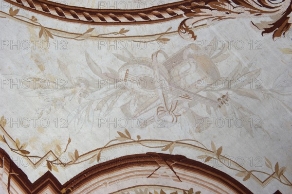 A so-called ceiling tapestry, tapestries, restitution provenance, fabrics, tapestries, knotted monograms, floral motifs, interior furnishings, weaving. In the center, a rosette surrounded by a floral wreath. Surrounded by rectangular frames with rounded shorter sides and floral garlands. In the corners are medallions with a knotted CKJ monogram. The border varies in color with plant patterns imitating the damask technique. Colors: pink, red, light brown, and olive. Early 19th century, France, fabrics, height 681 cm, width 385 cm., A patterned surface with a pale beige background showing a faint central decorative motif of leaves and a ribbon-like shape in very light tan and off-white hues, surrounded by a darker border composed of curved bands in shades of brown and tan; several vine-like lines with small leaves in light brown and gold run along the upper and lower edges, and there are scattered light tan spots and subtle fabric texture visible across the whole image.