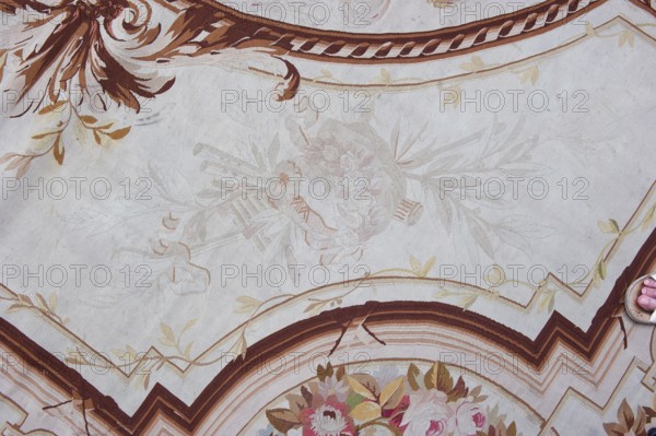 A so-called ceiling tapestry, tapestries, restitution provenance, fabrics, tapestries, knotted monograms, floral motifs, interior furnishings, weaving. In the center, a rosette surrounded by a floral wreath. Surrounded by rectangular frames with rounded shorter sides and floral garlands. In the corners are medallions with a knotted CKJ monogram. The border varies in color with plant patterns imitating the damask technique. Colors: pink, red, light brown, and olive. Early 19th century, France, fabrics, height 681 cm, width 385 cm., A patterned surface with a pale background showing ornamental flourishes in brown and tan tones, a decorative curved border with stripes in shades of brown and beige, and floral motifs in muted pinks, greens, and yellows; a few leafy vine designs in light tan and gold appear across the surface, and a partial view of a foot wearing a sandal is visible at the edge.