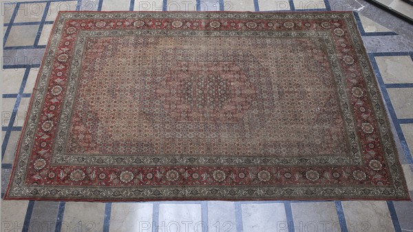 Carpet, In the center are four hexagonal medallions, one inside the other. Their interiors and the remaining small part of the background are filled with motifs of flowers, lanceolate leaves, and rosettes surrounding rhombuses. 3-band border with floral motifs. Materials: woolen knotted fabric. Colors: red in several tones, cream background, red, cream, and blue ornament. 2nd half of the 19th century, Iran. Function: interior furnishing/decorative tapestry. Dimensions: height 667 cm, width 415 cm., A large rectangular rug lies on a tiled floor, showing an intricate repeating pattern across its central field and a decorative border. The rug displays several colors including red, beige, brown, gray, and hints of green. The central area has a dense, small-scale geometric and floral motif in muted beige and brown tones with touches of red, while the border features a more pronounced red band with larger floral medallions in beige and brown framed by narrower bands in gray and beige. The su...