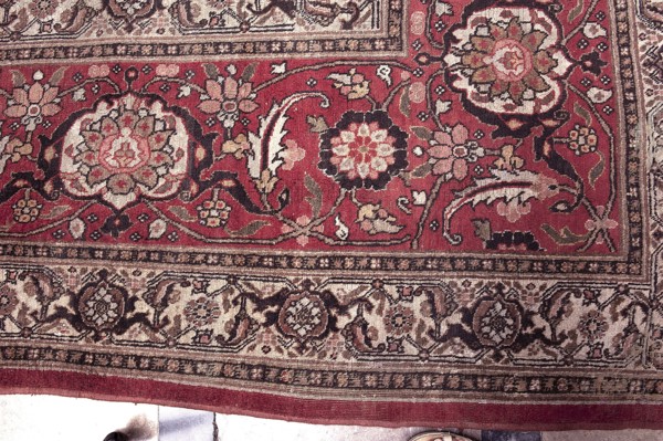 Carpet, Woolen knotted fabric, decorative tapestry, 2nd half of the 19th century, Iran. In the center are four nested hexagonal medallions. Their interiors and the remaining background are filled with motifs of flowers, lanceolate leaves, and rosettes surrounding rhombuses. Three-band border with floral motifs. Colors: several tones of red, cream background, blue ornament. Height 667 cm, width 415 cm., A patterned rug with a dominant deep red field and a border in beige and brown tones, featuring ornate floral motifs and curving vine-like shapes in shades of beige, cream, brown, black, olive green, and muted pink; a few large stylized flowers and medallion shapes appear within the red area, surrounded by smaller floral clusters and scrolling leaves, while the outer border shows repeating ornamental panels with intricate floral and arabesque designs; the rug edge is visible along the bottom with a narrow solid red band, and at the very bottom edge a small portion of a light-colored floo...