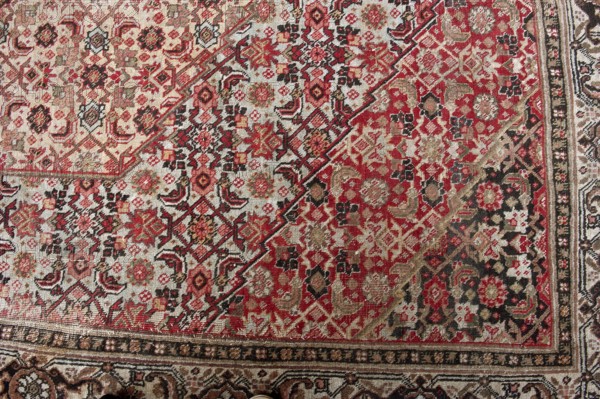 Carpet, Woolen knotted fabric, woven decorative carpet, 2nd half of the 19th century, Iran. In the center are four nested hexagonal medallions. Their interiors and the remaining background are filled with motifs of flowers, lanceolate leaves, and rosettes surrounding rhombuses. Three-band border with floral motifs. Colors: multiple tones of red, cream background, blue ornament. Height 667 cm, width 415 cm., A woven rug with an intricate geometric and floral pattern featuring a dominant mix of red and cream tones, accented by black, beige, brown, and hints of green. The design shows repeating medallions, star-like shapes, and small floral motifs arranged in diagonal bands and dense clusters, with a narrow ornamental border along one edge. The texture of the weave is visible, and the colors vary in intensity across the surface, creating areas that appear more saturated in red and others more muted in cream and beige. There is a subtle contrast between the darker black and brown accents a...