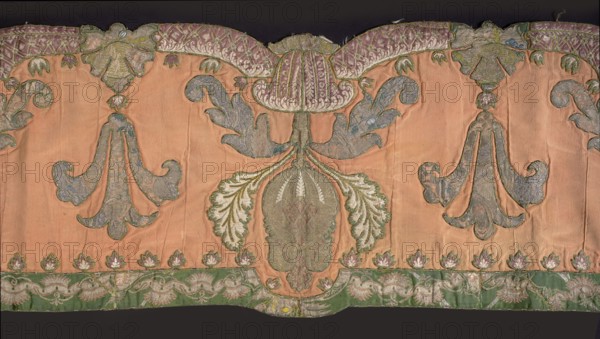 Lambrequin. Fragment of a lambrequin made of crimson silk grosgrain, sewn with fragments of patterned silk fabrics in green and brown. Decorated with palmette motifs, large serrated leaves, and small leaves arranged along the edges. The edges of the lambrequin are gently serrated. Silk lining. Early 18th century. Poland embroidery, France fabric. Materials: silk. Dimensions: height 41 cm, width 214 cm., Classification: decorative fabric / lambrequin., A decorative textile panel with a peach background featuring a central stylized floral motif in green and blue with pale green and white leaf shapes, flanked by two mirrored scrolling leaf designs in bluish-gray and green; a scalloped top border shows a pattern in purple and green, and a lower border band in green with pale green and white floral ornamentation; small stylized flower shapes in pink and white sit above the lower border; visible stitching and seams give a slightly wrinkled texture, and the overall palette includes peach, gre...