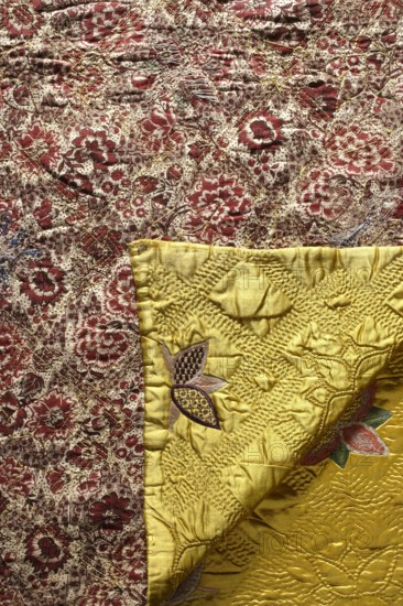 Quilt, Satin quilt, quilted and covered with multicolored embroidery in stylized flower patterns. Predominant colors: yellow background with pink, green, and navy blue ornament. Embroidery motifs include five-petal, star-like flowers. Quilted in rhombuses and repeating five-petal flower patterns. Lined with cotton fabric printed with floral patterns; burgundy and brown ornamentation on a cream background. 18th century, Europe. Materials: silk fabrics, printed and decorative fabrics, embroidery. Dimensions: height 205 cm, width 173 cm., A close-up of patterned fabric showing a textured floral textile in shades of reddish-brown and beige with a diagonal quilted pattern, alongside a folded piece of bright yellow fabric with stitched, raised embroidery forming floral and leaf motifs in colors including purple, pink, green, and brown; the image displays visible stitching, quilting textures, and slight wrinkles on the yellow fabric, with the reddish-brown and beige fabric featuring dense flo...