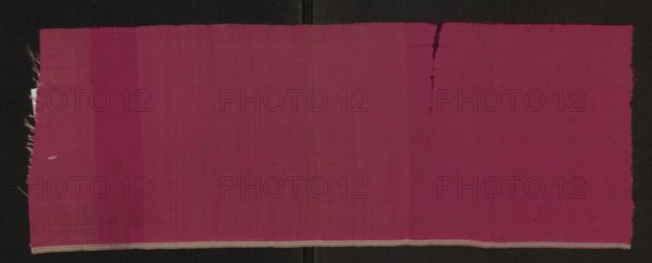 Fabric fragment, silk, 17th century, Europe. Rep weave with weft changed in zones creating lighter wide transverse stripes; weft changed from two-colored red–cream to single-colored red. Preserved selvedge; edge width with selvedge: 0.5 cm. Height 50 cm, width 18 cm., A large rectangular piece of woven fabric in shades of deep magenta and pink laid flat against a black background, showing subtle vertical variations in hue across its surface; the fabric has slightly frayed edges on the left and right sides and a small irregular tear or pull near the top right edge, with a narrow light-colored woven border along the bottom edge; the texture of the weave is visible, and there are a few faint darker smudges or marks on the fabric.