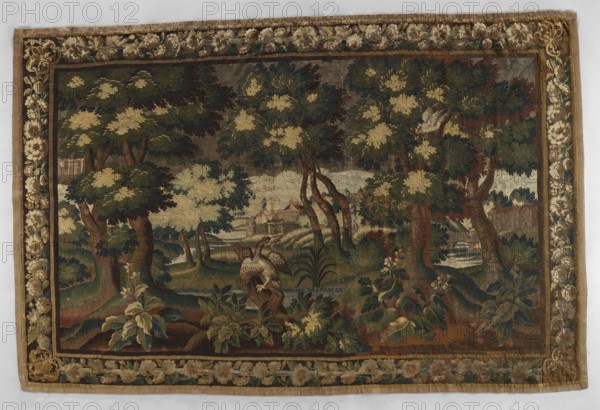 Tapestry — verdure;, In the center, on the trunk of a broken tree, a duck sits among clumps of sparsely growing trees. Palace buildings are visible through the trees; mountains in the distance. Border with floral motifs and flowers, floral ornaments, trees, landscapes, birds, decorative fabrics, interior decorations. Colors: brown, green, olive, and sand. 17th–18th century, Aubusson. Woven fabric; height 290 cm, width 437 cm., A woven scene shows a wooded landscape with a group of trees in the foreground and midground, their foliage in shades of green and pale yellow, and trunks in brown tones; a river or stream of pale blue weaves through the scene with a small bridge visible in the distance; the foreground includes a few large leafy plants in green and muted blue-green and a bird with outstretched wings perched on a stump rendered in brown, white, and gray; another bird appears perched higher in a tree with similar muted colors; a cluster of buildings with reddish roofs sits beyond t...