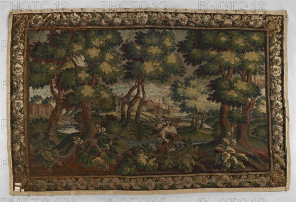 Tapestry — verdure. In the center, on the trunk of a broken tree, a duck sits among clumps of sparsely growing trees. Palace buildings are visible through the trees; mountains in the distance. Border with floral motifs and scattered flowers and birds. Materials: woven tapestry; colors: brown, green, olive, and sand. 17th–18th century, Aubusson. Function: decorative interior furnishing; height 290 cm, width 437 cm., A rectangular woven textile with a decorative border surrounds a central landscape scene featuring a wooded area with a group of trees, leafy canopies, and visible trunks and roots. In the foreground there are broad leaves and plants, and a perched bird with outstretched wings on a stump or branch. Behind the trees a body of water is visible, with more vegetation and a distant building partially obscured by foliage. The border has a repeating floral or circular motif. Dominant colors visible include green, brown, beige, tan, and touches of dark blue and muted red.