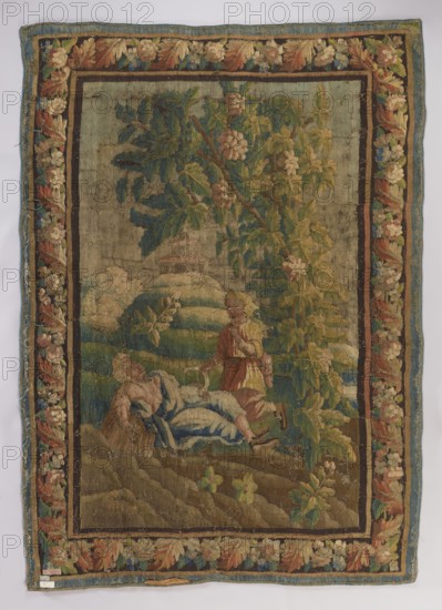 Figural tapestry depicting pastoral scenes with floral ornaments and borders. In the center foreground, under a tree, a sleeping girl lies with a sickle in her hand; above her a young boy kneels, raising a finger to his lips. Background shows a building with a double conical roof and a distant horizon. Border with floral motifs. Colors: brown, green, and sand. 2nd half of the 18th century, France. Woven fabrics; height 280 cm, width 195 cm., A rectangular woven tapestry with a wide floral border in shades of brown, beige, green, blue, and red surrounds a central pastoral scene; inside the border, a large tree with green and yellow leaves dominates the right side while branches with darker green leaves extend across the upper area. In the lower central area, a reclining figure dressed in blue and white striped fabric rests on the ground beside another seated or kneeling figure wearing garments in red and yellow tones, and a standing figure in a red and yellow outfit reaches toward them....