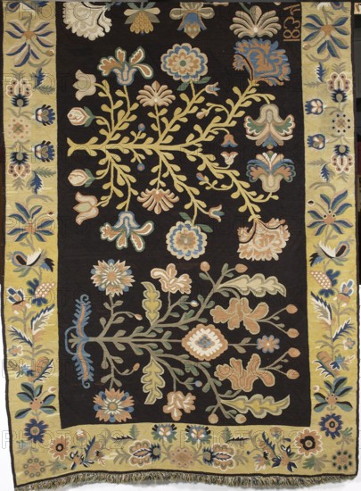 Court rug, Woven textile, fabrics, weaving; decorative interior furnishing with plant and floral motifs. Central field filled with motifs of four stylized trees with flowers and leaves—generally fanciful; carnation flowers are recognizable. Surrounded by a single-strand border with stylized flowers, fruit, and leaves arranged on a tendril. Colors: main field background black; borders sandy yellow; ornament in blue, greenish, dirty yellow, sapphire blue, and white. Woven signature and date at upper edge: JK1834, 1834. Place of origin: Poland. Dimensions: height 186 cm, width 416 cm., A rectangular textile with a dark central field decorated with an arrangement of stylized flowering branches and leaves in muted tones of yellow, beige, blue, green, and pale orange, surrounded by a lighter yellow border that repeats floral and leafy motifs in similar colors; the design includes a variety of rounded and scalloped flower shapes, tulip-like buds, curving stems, and layered leaves, with small ...