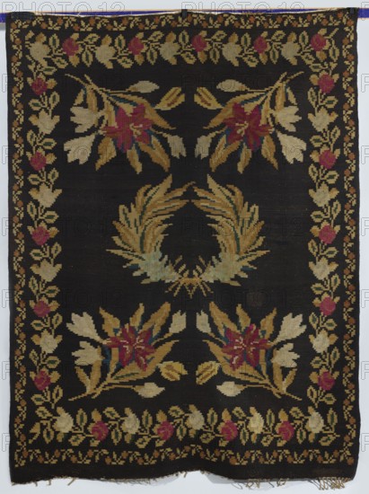 Kilim, In the center are motifs of two palm branches crossed at the base; in the corners are four branches with lily flowers. The kilim is surrounded by two floral tendril motifs. Colors: background dark brown; ornament variegated. Ca. 1850. Poland. Fabric. Height 201 cm; width 158 cm., A rectangular woven textile with a dark background featuring a central laurel-like wreath in shades of tan, gold, and muted green, surrounded by floral clusters in tan, gold, pink, red, and small accents of blue, with a decorative border of repeating floral motifs in similar tan, gold, pink, and red tones; the textile shows fringe at one short edge and the colors include black, tan, gold, pink, red, blue, and green.