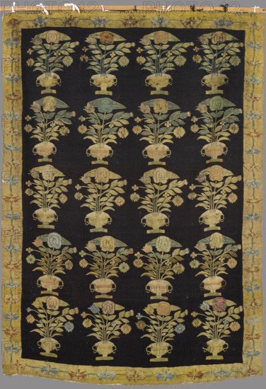 Kilim, Floral bouquet motifs placed in two-handled vases on a base, arranged in five horizontal rows of four motifs each across the main field. Surrounded by a narrow border with a floral tendril motif. Colors: black background in the central field, dirty yellow borders; ornamentation in pink, blue, green, cream. 19th century first half, Poland/Podolia Ukraine cultural region. Decorative fabric, manor and home furnishings; height 259 cm, width 185 cm., A rectangular textile with a dark background patterned in a repeating grid of floral arrangements in vases, each motif showing a vase holding stems, leaves, and rounded flower heads, all rendered in muted yellow, pale green, light blue, and soft pink tones; the central field is framed by a wide yellow border decorated with stylized floral shapes in blue, red, and beige; the overall colors visible include black, yellow, green, blue, pink, beige, and brown; the textile shows some areas of wear and slight color variation across the motifs a...