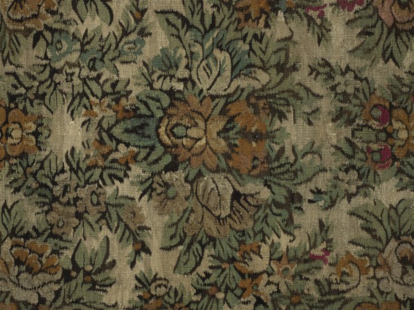 Court kilim. The main field is filled with flower and leaf motifs arranged symmetrically in repeating rhomboid arrangements. Surrounded by a three-stripe border: the middle, widest strip has a floral garland motif; the narrow outer strips feature floral and leafy tendrils. Colors: central field background off-white, borders tobacco-colored; ornamentation in green, magenta, tobacco, white, and black. Ca. 1850. Poland. Fabrics. Height 218 cm, width 215 cm., A patterned textile with an overall beige background featuring a repeating botanical motif in shades of green, muted teal, brown, tan, black, and small accents of magenta, showing stylized leaves and flowers in an ornate, symmetrical arrangement, with visible woven texture and areas of lighter and darker wear across the fabric.
