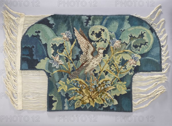 Tapestry coverings, repossession provenance. Decorative fabric for interior furnishings: a seat—bird with spread wings among large leaves and flowers; b backrest—flowers among large leaves. Colors: green, blue, yellow, ochre, and brown. 1st half of the 20th century, Vienna. Fabrics. Height 74 cm, width 109 cm., A woven textile with a central bird motif perched among plants, featuring a palette of teal, dark blue, green, beige, brown, yellow, and touches of pale pink and light blue; the bird has patterned wings in shades of beige and brown and stands amid leafy stems and floral elements rendered in yellow, green, and blue tones; the background shows abstract rounded shapes and flowing areas in teal, dark blue, and green; the textile has cream-colored fringe along both sides and a cream border on one edge, and the whole piece lies flat on a light gray surface.
