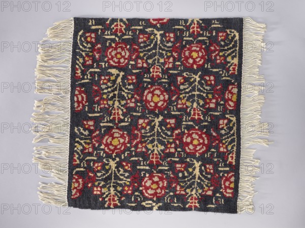 Tapestry covering; floral ornaments and stylized flower trees identical on the seat and backrest; fabrics and decorative borders; navy blue background with red, yellow, and cream ornamentation; interior furnishing; 1st half of the 20th century, Vienna; height 67 cm, width 74 cm., A rectangular woven textile with fringed edges lies flat on a pale surface; the central field is dark, almost charcoal, patterned with a repeating floral motif in red and muted yellow, with cream-colored curving vine shapes connecting the flowers, and a border of plain dark weave along the sides; the fringe at the shorter ends is off-white and loosely twisted, and the overall palette includes dark gray, red, yellow, cream, and a pale background.