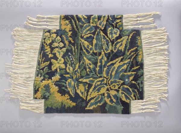 Tapestry covering, flowers, leaves, floral ornaments, fabrics, interior decorations, decorative fabrics, interior furnishings, furniture upholstery; a seat: forest landscape with a thicket of leaves; b landscape with a view of a mountain with a castle. Colors: green, blue and yellow. 1st half of the 20th century, Vienna. Fabrics. Height 72 cm, width 79 cm., A woven textile with a floral and leafy pattern in dark blue, deep green, teal, and mustard yellow tones, set against a pale gray background; the textile has a rectangular central panel with several overlapping leaf and flower shapes and a few stepped extensions at the top and bottom, and it is finished on both vertical edges with long cream-colored fringe strands, some of which are knotted.