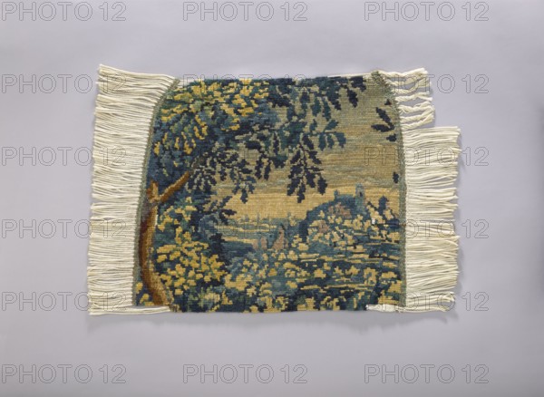 Tapestry covering, repossession provenance; flowers, leaves, floral ornaments; fabrics, interior decorations, decorative fabrics, interior furnishings, furniture upholstery; a seat, forest landscape with a thicket of leaves; b landscape with a view of a mountain with a castle. Colors: green, blue and yellow. 1st half of the 20th century, Vienna. Fabrics. Height 72 cm, width 79 cm., A rectangular woven textile with fringed edges lies flat against a pale gray background; the textile shows a landscape-like pattern in deep blue, muted yellow, and green tones with a visible tree trunk in brown and blue leaves, surrounded by dense foliage and textured horizontal bands, and cream-colored fringes extend from the shorter sides of the piece.