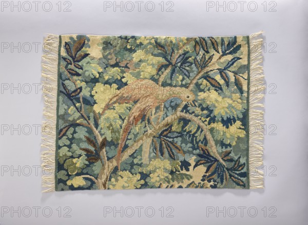 Tapestry chair cover, Repossession provenance. Design: leaves, floral ornaments, trees, birds, among thick foliage. Function: furniture upholstery/decorative fabric for interior furnishings. Materials: fabrics. Colors: yellow, green, blue, and pink. Date: 1st half of the 20th century. Place of origin: Vienna. Dimensions: height 65 cm, width 82 cm., A rectangular woven textile with fringe along the shorter edges lies flat against a pale gray background; the textile features a tapestry-like scene of plant forms and a perched bird, rendered in a palette of green, blue, yellow, brown, beige, and cream, with darker navy accents and touches of rust, showing leafy branches and layered foliage filling the composition.