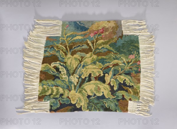 Tapestry chair cover, repossession provenance; design of flowers, leaves, and floral ornaments; decorative fabric for interior furnishings and upholstery gobelin-style tapestry; 1st half of the 20th century; Vienna place of origin; fabrics; height 68 cm, width 76 cm., A rectangular woven textile with cream-colored fringe along its shorter sides and parts of the longer sides, displaying a tapestry-like botanical design of large leafy forms and stems. The visible colors include various shades of green, teal, olive, brown, beige, blue, pink, and cream. The woven surface shows layered leaves with darker and lighter tones creating depth, and small areas of pink suggesting floral accents. The background areas transition between teal and brown tones, and the fringe is arranged in loose twisted strands against a light gray surface.