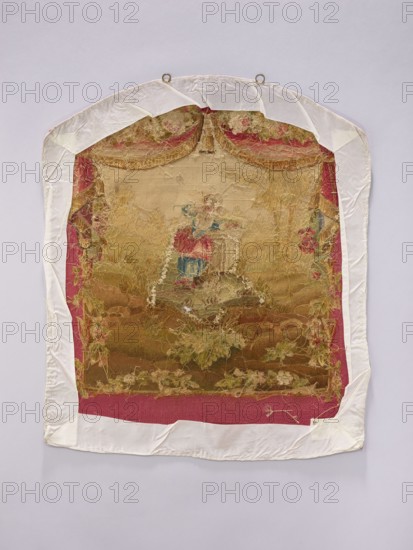 Tapestry covering, creator unknown, provenance, depicts flowers, leaves, floral ornaments, girls, pastoral scenes, boys; used as fabrics, interior decorations, interior furnishings, tapestries, furniture upholstery; with hanging draperies at the top and flowers around. Colors: beige, cream, green, sky blue, ochre, and red. Post–mid-18th century; possibly Beauvais or Aubusson origin. Height 71 cm, width 58 cm., A framed textile panel with a worn embroidered scene mounted on a pale background, showing a central figure in a dress with visible red and blue areas, surrounded by faded gold and beige embroidery and decorative floral motifs in shades of green and pink; the panel is bordered by a deep pink fabric and a white protective fabric wrap with metal hanging rings at the top, and the overall palette includes white, pink, red, blue, gold, beige, green, and brown.