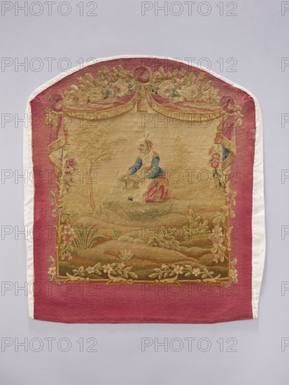 Tapestry covering, provenance, depicts flowers, floral ornaments, pastoral scenes, boys; fabrics used for interior decorations, furniture upholstery and hanging draperies with flowers around the top. Colors: beige, cream, ochre, green, blue, and red. Post–mid-18th century; possibly Aubusson or Beauvais. Materials: fabric. Height 71 cm, width 58 cm., A rectangular textile with a rounded top bordered by a wide pink band and narrow white edges surrounds a central woven scene in muted beige and tan tones; the scene shows a person wearing a blue top and pink skirt standing in a pastoral landscape with grassy mounds and scattered plants, framed at the top by a decorative swag of floral garlands in shades of green and pale yellow and flanked by ornate vertical floral borders with pink and green accents, while the lower border features a garland of pale flowers against the pink background.