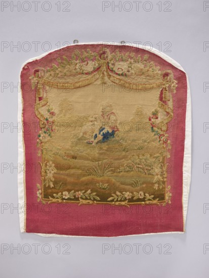 Tapestry covering, provenance, subject: flowers, floral ornaments, dogs, boys, pastoral scenes; function: interior furnishing/tapestry for furniture upholstery; scene: on grass with draperies suspended above and flowers around. Colors: beige, cream, green, ochre, blue, and red. Date: post–mid-18th century. Possible origin: Aubusson or Beauvais. Material: fabrics. Dimensions: height 71 cm, width 59 cm., A rectangular woven textile with a rounded top edge is displayed against a pale gray background, featuring a central scene in muted beige and olive tones framed by a wide deep pink border; the central area shows a pastoral motif with a figure in pale colors and blue garments seated on grassy earth in shades of green and brown, surrounded by floral and leafy embellishments in soft greens, pinks, and cream, and topped by a decorative swag and ribbon-like elements in beige and gold tones; the pink border contains floral corner decorations in green and light pink, and the textile is backed a...