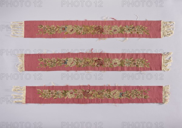 Six tapestry strips, creator unknown, origin, floral ornaments on fabrics for interior decoration; dark pink background with light pink, dark burgundy and olive ornaments; early 20th century 1st half, Vienna; length 102 cm, width 13 cm., Three long narrow woven fabric strips laid horizontally against a pale gray background, each strip in a dusty rose color with frayed cream-colored fringes at the ends. Each strip features a central floral band woven in muted greens, soft yellows, pale blues, and subdued browns, with clusters of stylized flowers and leaves forming a repeating motif. The rose-colored ground shows slight wear and loose threads along the edges, and the cream fringe appears twisted and uneven. The overall palette includes dusty rose, cream, green, yellow, blue, and brown.