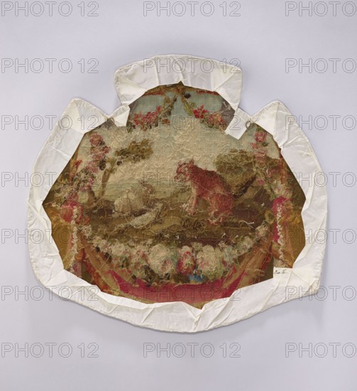 Tapestry armchair seat cover, unknown provenance. Design: flowers, floral ornaments, hares, foxes. Materials/subject: fabrics, decorative fabrics, tapestry upholstery/draperies with flowers around. Colors: beige, brown, ochre, pink, red, blue. Date: post–mid-18th century. Possible origin: Aubusson, or Beauvais . Dimensions: height 58 cm, width 61 cm., A worn circular textile with frayed edges is mounted on a pale background and partly covered by a white fabric border that is folded and pinned at several points; the central textile shows a faded scene with muted browns and beiges forming a landscape, a reddish figure against greens and olive tones, and areas of pink and rose-colored flowers, with touches of blue and cream among tangled threads and worn fibers.