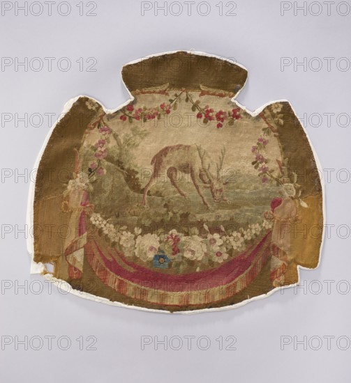 Tapestry covering, provenance, depiction of flowers, floral ornaments, and a deer; decorative fabric for interior furnishings, upholstery or drapery suspended from below with flowers surrounding. Colors: beige, brown, pink, red, and white. Post–mid-18th century. Possibly Beauvais or Aubusson. Fabric. Height 57 cm, width 62 cm., A worn, irregularly shaped textile with a central circular scene showing an animal with slender legs and antlers bending down amid a muted landscape, surrounded by garlands of flowers and foliage; the textile features draped ribbon-like swags at the lower edge and decorative tassel-like elements at the sides. Visible colors include shades of beige and brown in the background, green in the foliage, pink and red in the flowers and swags, cream and off-white in the floral garlands, and small accents of blue. The textile shows frayed edges and areas of wear and discoloration, mounted against a light gray background.