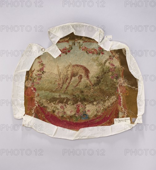 Tapestry covering, provenance, depiction of flowers, floral ornaments, and a deer. Decorative fabric intended for interior furnishings—furniture upholstery, tapestries, draperies suspended from below with surrounding flowers. Colors: beige, brown, pink, red, and white. Post–mid-18th century. Possible places of origin: Beauvais or Aubusson. Fabric; height 57 cm, width 62 cm., An oval textile fragment with frayed edges is mounted on a pale background and partially wrapped with off-white fabric tape; the textile shows a central animal figure bending downward surrounded by floral and foliage motifs, with colors including muted brown, beige, faded green, dusty pink, deep red, and small areas of blue, and the mounted piece displays loose threads and worn areas across its surface.