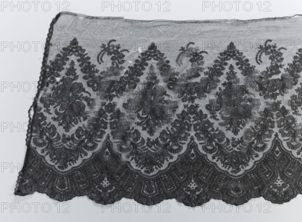 Crinoline ruffle, clothing accessories, garlands ornament, Chantilly lace, floral ornaments, lace, fashion. Bouquets placed in medallions created with garlands above and below them. Below the lower garland, a fan-shaped lace motif. The lower edge is serrated, the upper one straight. 2nd half of the 19th century, Chantilly place of origin, clothing, lace, width 54 cm, length 440 cm, A section of patterned lace fabric laid flat, showing an intricate floral and scalloped design in dark gray and lighter gray tones; the lace has a sheer background with denser embroidered floral motifs and swirling leaf shapes, scalloped edges along the bottom with repeating fan-like arches, and a slightly uneven top edge; a few loose threads are visible along the sides, and the overall color palette is shades of gray against a pale gray background.