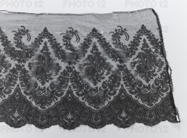 Crinoline ruffle;, clothing accessory; garlands ornament; Chantilly lace with floral ornaments; lace; fashion. Description: Bouquets placed in medallions created with garlands above and below them. Below the lower garland, a fan-shaped lace motif. Lower edge serrated, upper edge straight. 2nd half of the 19th century; Chantilly place of origin. Clothing lace. Dimensions: width 54 cm, length 440 cm., A section of delicate lace fabric is shown against a pale background, featuring an ornate floral and scalloped pattern; the lace displays a repeating arrangement of large floral motifs surrounded by smaller blossoms and leafy scrollwork, framed by a scalloped lower edge with intricate geometric and floral embroidery. The texture appears sheer and fine with visible mesh between the embroidered elements, and the edges show slight irregularities and fraying. The visible colors include shades of dark gray for the lace and light gray to off-white for the background.