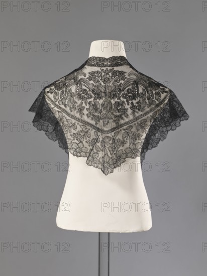 Mantilla, floral ornaments; Chantilly lace; lace; clothing accessory; women's clothing; fashion; mantilla, triangular with highly raised upper corners; 2nd half of the 19th century; France; lace; height 36 cm, A white mannequin torso displays a delicate black lace shawl draped over the shoulders, the lace featuring an intricate floral and leaf pattern with scalloped edges and varying densities of mesh; the background is a smooth gray, and the mannequin stand is a darker gray metal.