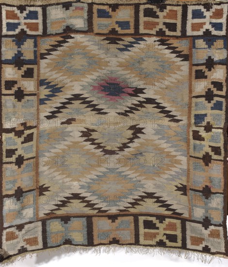 Kilim, Kilim, Podolia Ukraine origin; Poland cultural association. Decorative fabric for interior design, 1st half of the 19th century. Hand-stitched kilim with central field filled with rhombus motifs with stepped contours and slight overlapping in the center. Surrounded by a border of rectangle-like patterns with triangular notches on the longer sides. Geometric ornament. Colors: dirty gray, light blue and gray-blue, brown, ochre, brick red, with some navy blue and dark pink. Materials: fabrics. Height 141 cm; width 131 cm., A woven textile with a central field of zigzag and diamond motifs in muted beige, tan, light blue, pale gray, dark brown, and a small area of muted pink, surrounded by a geometric border of interlocking rectangular and plus-shaped motifs in beige, tan, blue, cream, and dark brown; the textile shows frayed edges and loose fringe at one end and variations in pile and wear across its surface.