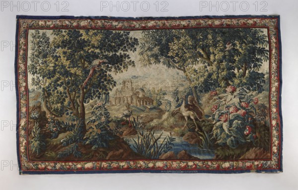 Tapestry-verdure, In the foreground, a river with overgrown banks and two large birds on opposite banks. To the right and left, trees; in the distance, through the trees, ruins of an arcaded building. Border with floral motifs. Colors: sapphire, green, red, blue, and cream. The manufacturer's signature is woven in large letters on the border tape. 1st half of the 18th century, Aubusson origin. Fabric. Height 287 cm, width 484 cm., A woven scene framed by a decorative border showing a wooded landscape with a stream, rocks, and a ruined building in the distance, featuring a group of large leafy trees in green, blue, and yellow tones, several flowering plants with red and pink blooms, a few birds perched in the trees and near the water with feathers in shades of red, white, and gray, and a large bird with brown and white plumage standing near the stream; the border includes floral detailing in red, cream, blue, and green, and the overall palette includes various shades of blue, green, bro...