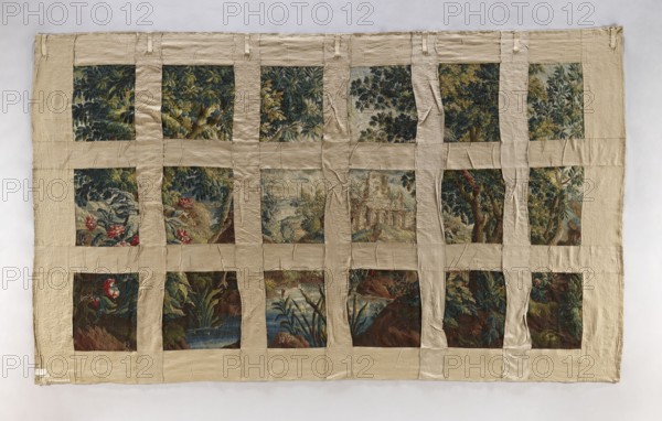 Tapestry-verdure, Floral ornaments, birds, trees, landscapes, fabrics, tapestries, interior furnishings, interior decorations, decorative fabrics. In the foreground, a river with overgrown banks and two large birds on opposite banks. To the right and left, trees. In the distance, through the trees, ruins of an arcaded building. Border with floral motifs. Colors: sapphire, green, red, blue, and cream. The manufacturer's signature is woven in large letters on the border tape. 1st half of the 18th century, Aubusson. Materials: fabrics. Height 287 cm, width 484 cm., A large rectangular textile composed of a grid of fabric panes framed by beige strips, showing a patchwork of painted or woven scenes with foliage, trees, water, rocks, and floral elements. The visible colors include beige framing, deep greens, dark blues, muted browns, touches of red, pale blue-gray, and hints of yellow and olive. Several panes display dense leafy canopies, a few show tree trunks and branches, a group depict w...