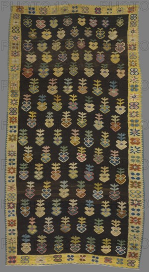 Kilim, kilim; decorative fabric with floral patterns; Poland; 18th–19th century. Kilim with motifs of stylized and geometrical flower branches arranged in 15 rows of 6 or 7 each, surrounded by a narrow border with floral rosette motifs. Colors: black background in the central part and yellow in the border; ornamentation in yellow, sand, light brown, green, pink, and blue. Materials: fabrics; height 285 cm, width 150 cm., A rectangular woven textile with a dark background covered in a repeating grid of stylized plant or flower motifs in a range of pastel and muted colors including yellow, pink, blue, green, beige, and brown, each motif consisting of a heart-like lower shape with a vertical stem and small branching elements above; a yellow border runs around the textile decorated with a sequence of small floral or geometric medallions in colors such as red, blue, green, pink, and beige, and the edges show variations in weave and slight irregularities.