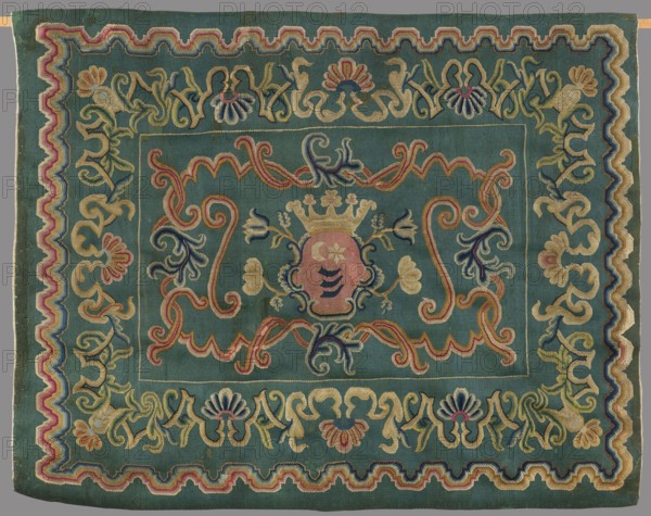 Kilim, In the center of the main field is a shield composed of ogee and rooster combs – the Wolda coat of arms, a variant of the Traby coat of arms. Above the shield is a large five-pronged crown. Motifs of two pairs of branches with large-cup flowers branch from the shield. Around the shield, filling the background, are symmetrically arranged motifs of ogee and acanthus leaves. The kilim is surrounded by a border decorated with fan-shaped palmettes, acanthus leaves, and wavy line segments. Colors: peacock-green background; ornamentation in sand, pink, yellow, and navy blue. Second half of the 18th century, Poland. Fabric. Height 150 cm; width 185 cm., A rectangular textile with a teal background featuring an ornate embroidered design in colors including gold, pink, blue, cream, and green; the central area shows a stylized crest-like shape with a crown motif above it, surrounded by flowing scrolls and floral elements in pink, gold, navy blue, and cream; a decorative inner border frames...