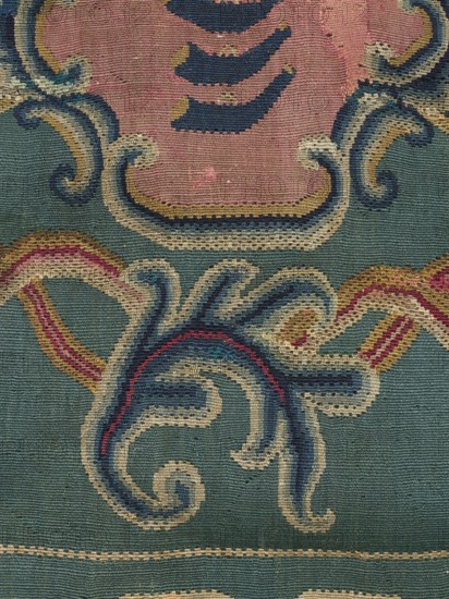 Kilim, In the center of the main field is a shield composed of ogee and rooster comb motifs — the Wolda coat of arms, a variant of the Traby coat of arms. Above the shield is a large five-pronged crown. Motifs of two pairs of branches with large-cup flowers branch from the shield. Around the shield — filling the entire background — are symmetrically arranged motifs of ogee and acanthus leaves. The kilim is surrounded by a border decorated with fan-shaped palmettes, acanthus leaves, and wavy line segments. Colors: peacock-green background; ornamentation in sand, pink, yellow, and navy blue. 2nd half of the 18th century, Poland. Woven fabric kilim, height 150 cm, width 185 cm., A close-up of a woven textile showing an ornate pattern with flowing, curling shapes and curved bands. The background is a muted teal green, with large areas of dusty rose and accents in navy blue, cream, mustard yellow, deep red, and pink. The design features scalloped and wave-like motifs outlined by contrasting...