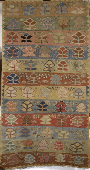 Kilim, kilim; interior design decorative fabric; Poland culture; mid-19th century, Podolia; woven flatweave measuring 226 cm high × 120 cm wide. The surface is divided into twelve horizontal stripes of varying colors. Within the stripes are arranged twigs—palmettes five in each. No border; only a serrated pattern runs around the edges. Colors: brown, tobacco, pink, and light blue., A vertical woven textile with repeating stylized leaf or tree motifs arranged in horizontal bands, featuring a palette of earthy and muted colors including beige, tan, brown, rust, green, blue, navy, red, and pale yellow; the motifs are outlined in contrasting threads and vary in fill color across rows, with several small elongated oval shapes interspersed between the motifs and occasional textured or patterned background bands in shades of blue, green, and rust; the textile edges show a zigzag decorative border and a fringe at the bottom, and the overall surface displays woven texture and slight irregularit...