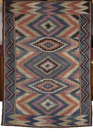 Kilim, Kilim with vertical stripes in rhombus motifs that merge concentrically into hexagons, their contours formed by stepped lines. Borders: shorter sides wide with overlapping stepped-line motif shaped like the letter M; longer sides narrower with a zigzag-line motif. Geometric ornament; colors dark blue, white, brick red, and pink. Late 19th century, Poland. Fabrics. Height 178 cm, width 123 cm., A rectangular woven textile with a repeating geometric diamond and zigzag pattern in colors including blue, pink, red, cream, black, and beige; the design features a central vertical series of large diamond motifs surrounded by stepped chevrons and smaller angular shapes, flanked by vertical border bands with a stair-step motif, and top and bottom edges that show alternating elongated chevron forms; the overall surface shows tightly woven texture and symmetrical, angular motifs creating a layered, multicolored appearance.