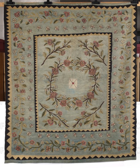 Kilim, kilim, court kilim, fabrics. In the center of the main field is a motif of a sealed envelope surrounded by a wreath woven with roses and small four-petal flowers and leaves. In the corners are motifs of twigs with flowers and rose buds. The kilim is surrounded by a wide border with a motif of a winding branch with bellflowers and six-petal flowers. This wide border is bordered on both sides by narrow zigzag trails. Colors: blue, white, pink, green, straw, ochre, and black. 1st half of the 19th century, Poland. Fabrics, height 206 cm, width 165 cm., A rectangular textile with a pale blue-green background features a central panel framed by a narrow dark border and a zigzag patterned band in dark and beige; inside the central panel there is a circular floral wreath of stylized roses and small flowers connected by curving branches and leaves in shades of pink, green, olive, brown, beige, and cream, with a small diamond-shaped motif at the center; surrounding the central panel is a w...