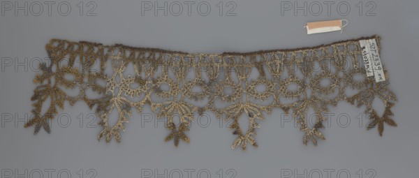 Lace, bobbin lace, 16th–17th century, Flanders, fabric, width 7 cm, length 27 cm;, A delicate lacy trim with an openwork pattern lies flat against a gray background, showing an ornate repeating design of loops, floral shapes, and dangling leaf-like motifs; the lace appears in shades of metallic gold and darker brown, with subtle variations creating a textured, slightly aged look, and a small fabric tag in beige and white is attached near one end.