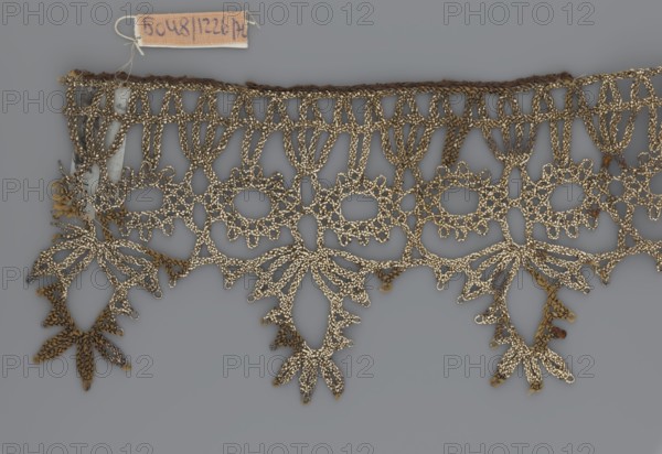 Lace, bobbin lace, 16th/17th century, Flanders, fabric, width 7 cm, length 27 cm, A length of decorative lace with an openwork floral and geometric pattern in metallic gold and brown tones laid against a neutral gray background, showing scalloped lower edges with dangling petal-like motifs, interconnected loops and oval openings, braided upper edge, and small fabric remnants attached at one end in muted beige and gray.