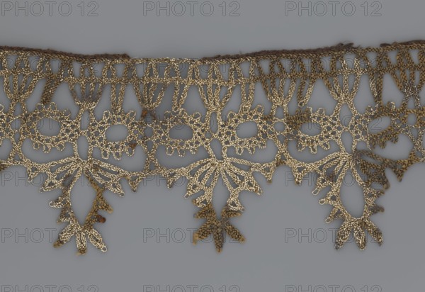 Lace, bobbin lace, 16th/17th century, Flanders, fabric, width 7 cm, length 27 cm, A decorative strip of openwork lace with an intricate repeating pattern lies against a neutral gray background; the lace shows a scalloped top edge and a row of looped motifs transitioning into a lower row of fan- and petal-shaped drops with pointed tips, and a few dangling floral-like pendants; the material appears textured with a metallic sheen in shades of gold and pale beige, with some areas showing darker brown and muted ochre tones.