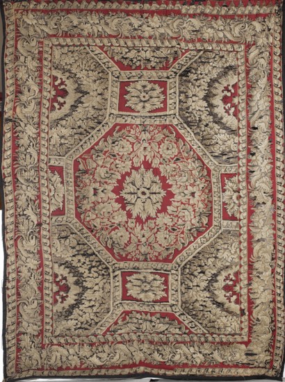 Kilim, In the center a leafy rosette is surrounded by a floral wreath and an octagonal frame. Geometric figures extending from the edges of the central octagon—rectangles, half-rectangles, half-octagons, and their fragments—are filled with similar floral ornamentation. The fabric is surrounded by a three-stripe border: the central, widest strip features a dense floral garland motif, while the two narrower strips feature writhing ribbon motifs. Materials: fabrics. Colors: brown, green, white, and red. Date: c. 1830. Place of origin: Milowidy. Dimensions: height 255 cm, width 195 cm. Genres/uses: interior furnishings, decorative fabrics, manor and home furnishings, court kilims., A large rectangular woven textile with an ornate symmetrical floral design, featuring a dominant red background and extensive beige and black detailing; a central octagonal medallion filled with beige floral motifs sits on the red field, surrounded by several framed panels with beige leafy patterns and areas of ...