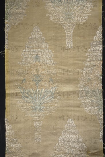 Three fragments of a chasuble, fabrics, silk, patterned, Persian Persia, plant patterns. Three light-yellow fabric fragments parts of a chasuble brocaded with motifs of cypress trees and filled with a stylized lily. Pale yellow grosgrain background; pattern brocaded with metal thread and silk yarn in green, celadon, pink, and orange. 18th century, Iran; dimensions reported as 49 x 16?, height 95 cm, width 24 cm., A vertical piece of fabric with a muted beige background patterned with a repeating ornate motif resembling stylized floral or tree forms in pale silver, light blue, and soft peach tones; the motifs have textured woven details and are arranged with several partial motifs visible at the edges, and the fabric shows some irregularities such as loose threads and slight creases along its surface with a thin darker border visible along the left and right edges.