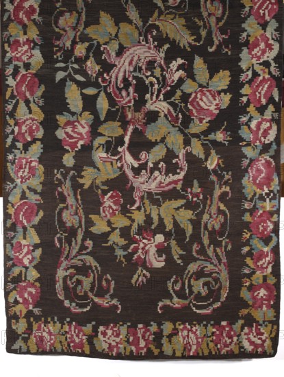Kilim, court kilim so-called Stanislawowski, mid-19th century. Woven decorative fabric kilim from Poland, height 305 cm, width 175 cm. In the center is an inverted S motif made of acanthus leaves entwined with flowers and leaves; S-shaped acanthus motifs appear in the corners. The field is surrounded by a tendril with large rose flowers and leaves. Colors: background brown; ornament in pink, red, green, light blue, and yellow., A woven textile with a dark brown background featuring an overall floral and scrolling vine pattern in shades of pink, dusty rose, mustard yellow, pale blue, and cream, with a border of repeating floral motifs along the edges and a lower edge band of clustered floral shapes, displaying stylized leaves and rounded flower forms arranged symmetrically across the surface.