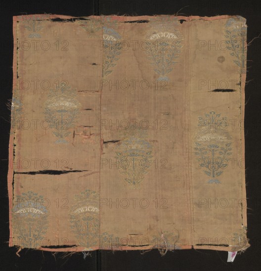 Fabric fragment, fabrics, origin: Persia Iran, 18th century. Fragment of silk patterned fabric with preserved selvedge width 0.1. Design: flower trees growing from stylized trapezoidal mound-roots, topped with flower branches growing from a plume made of stylized floral calyces. Colors: pink background with a grosgrain weave; brocades in grass-green, blue, steel-grey, salmon, and white. Dimensions: height 43 cm, width 43 cm., A rectangular piece of worn fabric laid flat with frayed edges and several horizontal tears, showing a faded tan background with a repeating floral medallion pattern in muted blue and pale gold, the medallions arranged in vertical columns and appearing at intervals across the surface; the fabric has a narrow faded pink border along the sides and bottom, and there are areas of darker discoloration and small spots, with some loose threads and uneven stitching visible along the edges.