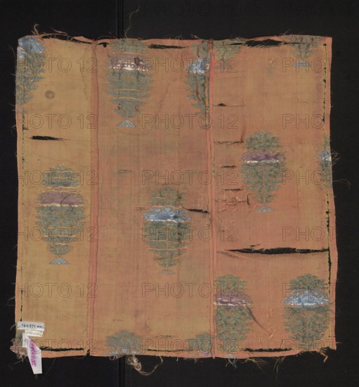 Fabric fragment, silk, origin: Persia Iran, 18th century. Fragment of patterned silk fabric with preserved selvedge width 0.1. Design: flower trees growing from stylized trapezoidal mound-roots, topped with flower branches growing from a plume made of stylized floral calyces. Colors: pink background with grosgrain weave; brocades in grass-green, blue, steel-grey, salmon, and white. Dimensions: 43 cm x 43 cm., A rectangular piece of worn fabric displayed on a dark background, showing a faded warm peachy-pink base with vertical seams creating panel divisions, frayed edges and several horizontal tears and holes; decorative motifs of stylized floral or tree-like shapes appear repeatedly across the surface in muted green, pale blue, soft purple, and a touch of metallic sheen, with some motifs partially damaged or missing; overall texture appears thin and aged with loose threads and slight discoloration in places.