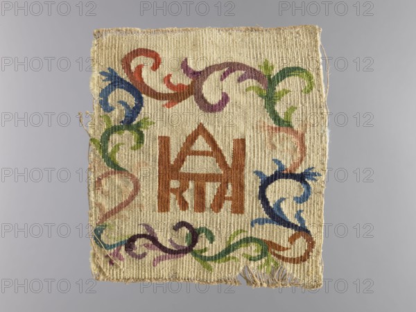 Palka?;, church paraments; unknown origin; plant ornaments; sacred art; churches and chapels furnishings; 18th century; liturgical parament; height 21 cm; width 19.5 cm, A rectangular woven textile with a beige background displaying a central geometric motif in brown, surrounded by a decorative scrolling border in shades of green, blue, purple, and reddish-brown; the fabric has a textured, ribbed weave and slightly frayed edges with loose threads, and the image background is a neutral light gray.
